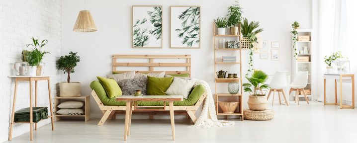 Wooden table in front of green couch with cushions in floral living room interior with leaves posters and suitcase on shelf